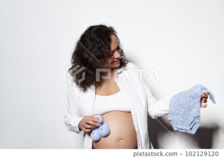 Curly haired young adult pregnant woman with big belly, holding blue bodysuit and knitted baby booties for future newborn, preparing things in pregnancy time, smiling, isolated on white background Curly haired young adult pregnant woman with big belly, holding blue bodysuit and knitted baby booties for future newborn, preparing things in pregnancy time, smiling, isolated on white background 102129120