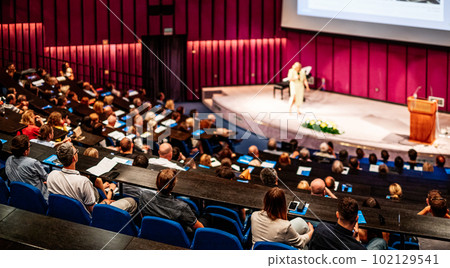 Business and entrepreneurship symposium. Female speaker giving a talk at business meeting. Audience in conference hall. Rear view of unrecognized participant in audience. 102129541