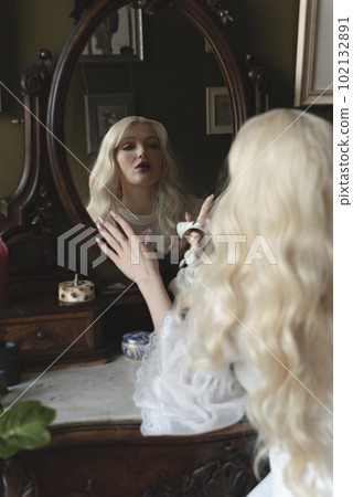 Beautiful young woman in a white dress sitting at the chair and reflecting in the mirror Beautiful young woman in a white dress sitting at the chair and reflecting in the mirror 102132891