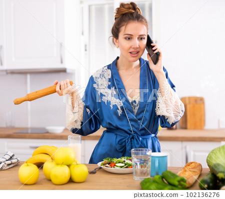 Dissatisfied woman with rolling pin in hands talking on phone in kichen Dissatisfied woman with rolling pin in hands talking on phone in kichen 102136276