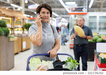 Mature woman talking on phone in grocery store with groceries in cart Mature woman talking on phone in grocery store with groceries in cart 102136282