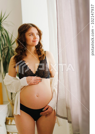a beautiful pregnant woman in a black underwea and white shirt by the window 102147937