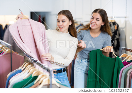 Positive young women testing fabrics of green and pink cardigans while shopping in a showroom Positive young women testing fabrics of green and pink cardigans while shopping in a showroom 102152867