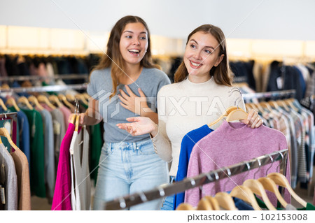 Happy young women holding winter jumpers and sweaters on racks during shopping in a boutique 102153008