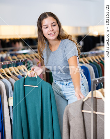 Positive young woman is thinking about buying a turquoise cardigan while walking through a clothing store Positive young woman is thinking about buying a turquoise cardigan while walking through a clothing store 102153322