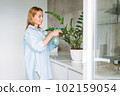 Young woman in blue shirt with spray with water in hands takes care of houseplant in kitchen at home 102159054