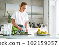 Young woman in white t-shirt and blue jeans cooking healthy food with cucumber in kitchen at home 102159057