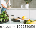Fresh green healthy fruits and vegetables on table while woman cooking smoothie in kitchen at home 102159059