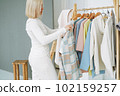 Young blonde woman stylist seller chooses clothes on hanger in bright studio 102159257