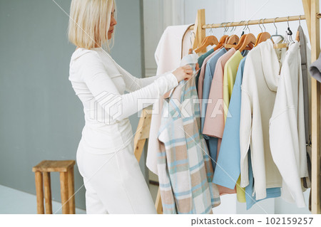 Young blonde woman stylist seller chooses clothes on hanger in bright studio Young blonde woman stylist seller chooses clothes on hanger in bright studio 102159257