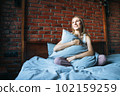 Young adult smiling woman with blonde long hair in casual sitting on bed with pillow at the home 102159259