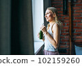 Young adult woman with blonde long hair in casual with green smoothie near window at the home 102159260