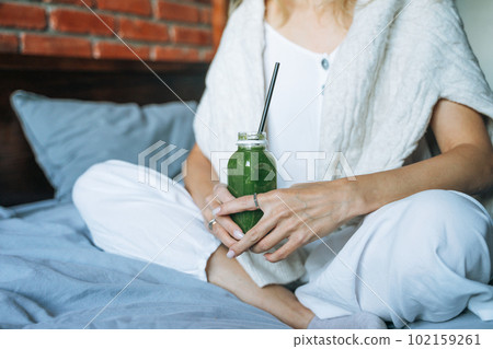 Crop photo of Young woman in casual clothes with green smoothie sitting on bed at the home Crop photo of Young woman in casual clothes with green smoothie sitting on bed at the home 102159261