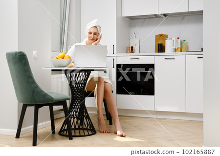 Young woman in bathrobe sits in the kitchen and browsing social media on laptop Young woman in bathrobe sits in the kitchen and browsing social media on laptop 102165887