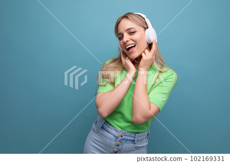 cute blond girl in casual outfit listens to music on the app in big white headphones on a blue cute blond girl in casual outfit listens to music on the app in big white headphones on a blue 102169331