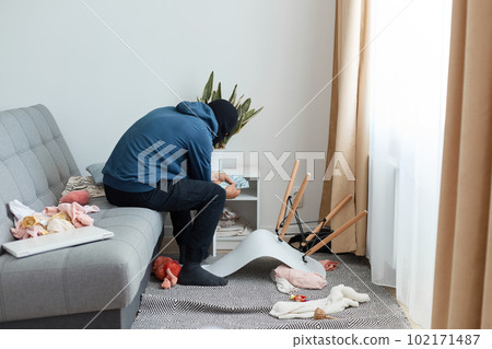 Man robber wearing blue hoodie and black balaclava, the thief found money in the nightstand in the house which trying to rob, sits on the couch and counts the cash he is holding in his hands. Man robber wearing blue hoodie and black balaclava, the thief found money in the nightstand in the house which trying to rob, sits on the couch and counts the cash he is holding in his hands. 102171487
