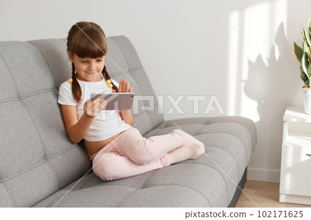 Adorable little cute dark haired female child wearing white casual t shirt using mobile phone sitting on couch, watching cartoons or playing video game, searching interesting children content. Adorable little cute dark haired female child wearing white casual t shirt using mobile phone sitting on couch, watching cartoons or playing video game, searching interesting children content. 102171625
