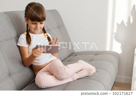 Indoor shot of little cute dark haired female child wearing white casual t shirt using cell phone sitting on couch, looking at mobile phone display and showing v sign. Indoor shot of little cute dark haired female child wearing white casual t shirt using cell phone sitting on couch, looking at mobile phone display and showing v sign. 102171627