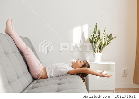Side view of cute dark haired female child wearing casual style attire lying on bed with raised legs and laughing, stretching arms, spending time alone, having fun at home. 102171651