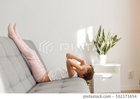 Sleepy adorable child lying on gray comfortable sofa with raised legs and rubbing her eyes, feels tired, wants sleeping , needs time to rest, wearing casual style clothing. 102171654