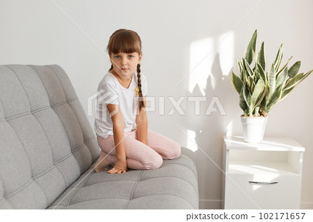 Indoor shot of adorable sad female kid with dark hair and pigtails sitting on couch and looking at camera with upset facial expression, wearing white casual style t shirt. Indoor shot of adorable sad female kid with dark hair and pigtails sitting on couch and looking at camera with upset facial expression, wearing white casual style t shirt. 102171657