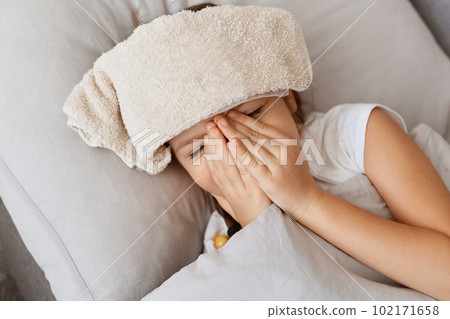 Closeup of sick little baby girl with fever sleeping in bed, low temperature with wet towel on forehead, covering face with palms, preschooler kid having terrible headache. Closeup of sick little baby girl with fever sleeping in bed, low temperature with wet towel on forehead, covering face with palms, preschooler kid having terrible headache. 102171658