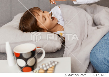 Side view of little unhealthy girl lying on cough, mother or doctor taking care of her sick child, giving medicine, treating influenza, drinking syrup from coughing. 102171668