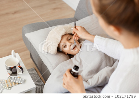 Horizontal shot of little sick girl lying on sofa, mother giving her spoon with syrup, family posing in light room, kid taking medicine from coughing or high temperature. Horizontal shot of little sick girl lying on sofa, mother giving her spoon with syrup, family posing in light room, kid taking medicine from coughing or high temperature. 102171789