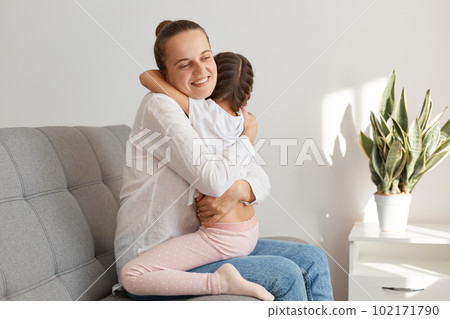 Charming female child hugging her lovely mother, people embracing each other and expressing love and care, posing at home, sitting in living room on sofa. Charming female child hugging her lovely mother, people embracing each other and expressing love and care, posing at home, sitting in living room on sofa. 102171790