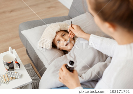 Portrait of little sick girl with wet towel on her forehead lying on sofa, mother giving her spoon with syrup, treating her lovely child from influenza or virus. Portrait of little sick girl with wet towel on her forehead lying on sofa, mother giving her spoon with syrup, treating her lovely child from influenza or virus. 102171791