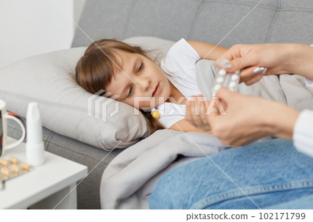 Portrait of unhealthy little girl lying on sofa under blanket, faceless mother holding pills, child suffering from fever, influenza symptoms or sore throat, home treatment. 102171799