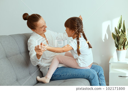 Indoor shot of happy smiling woman wearing white shirt and jeans, sitting on sofa with her charming daughter, family playing together, holding each others hands. Indoor shot of happy smiling woman wearing white shirt and jeans, sitting on sofa with her charming daughter, family playing together, holding each others hands. 102171800