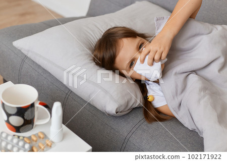 Image of unhealthy sick little girl lying on sofa under blanket and suffering runny nose, using tissue, looking nat camera, having bad mood, catching cold. Image of unhealthy sick little girl lying on sofa under blanket and suffering runny nose, using tissue, looking nat camera, having bad mood, catching cold. 102171922