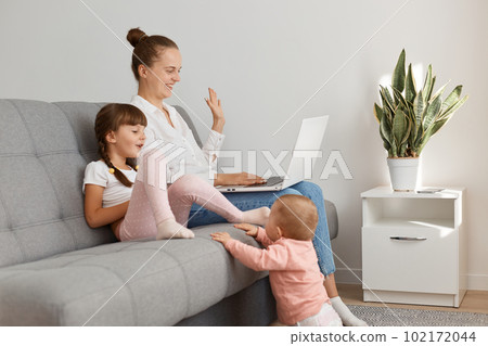 Indoor shot of optimistic smiling young woman with her little daughter with pigtails, having video call, waving hand to camera, expressing positive emotions. 102172044