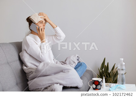 Unhealthy woman with bun hairstyle wearing white sweater sitting on cough, holding cell phone in hand and talking with doctor, keeping hand with towel on her forehead, having high temperature. Unhealthy woman with bun hairstyle wearing white sweater sitting on cough, holding cell phone in hand and talking with doctor, keeping hand with towel on her forehead, having high temperature. 102172462