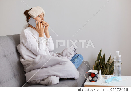 Image of sick young adult woman sitting on cough wrapped in blanket, posing with wet towel on her head, talking via mobile phone, suffering sore throat, influenza symptoms. Image of sick young adult woman sitting on cough wrapped in blanket, posing with wet towel on her head, talking via mobile phone, suffering sore throat, influenza symptoms. 102172466