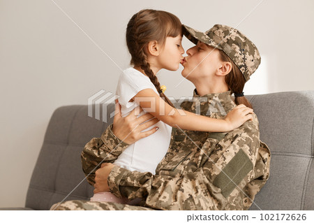 Indoor shot of happy military woman wearing camouflage uniform and cap, returning home from war or army, sitting on sofa with her daughter, kissing her child, missing, hugging each other. 102172626