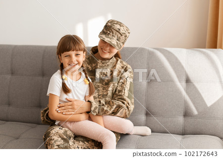 Image of smiling dark haired female kid with braids and her mother sitting on sofa a t home, woman wearing camouflage uniform and cap posing with her daughter. Image of smiling dark haired female kid with braids and her mother sitting on sofa a t home, woman wearing camouflage uniform and cap posing with her daughter. 102172643