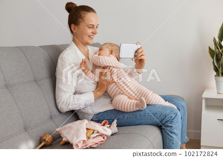 Indoor shot of smiling woman with hair bun wearing white shirt and jeans sitting on sofa with her infant daughter and having video call with her friend, spending time with child and talking. 102172824