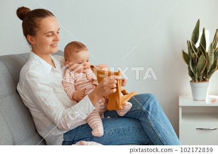 Indoor shot of young adult Caucasian mother sitting on sofa with her infant daughter and holding child's clothing, having positive facial expression, admires new attire. Indoor shot of young adult Caucasian mother sitting on sofa with her infant daughter and holding child's clothing, having positive facial expression, admires new attire. 102172839