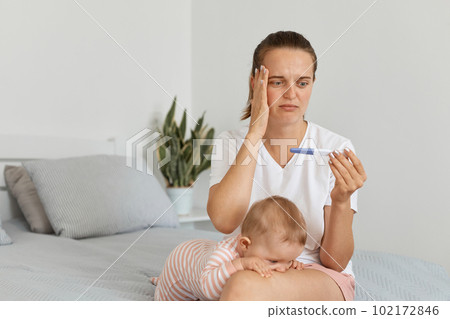 Indoor shot of sad young adult pregnant woman with pregnancy test, posing in her bedroom with toddler baby, wearing white t shirt, touching her head, having serious problems. Indoor shot of sad young adult pregnant woman with pregnancy test, posing in her bedroom with toddler baby, wearing white t shirt, touching her head, having serious problems. 102172846