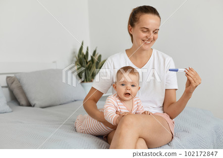Horizontal shot of attractive female with pigtail holding pregnancy test in hands, sitting on bed in bedroom with her infant daughter, looking at positive result and expressing happiness and joy. Horizontal shot of attractive female with pigtail holding pregnancy test in hands, sitting on bed in bedroom with her infant daughter, looking at positive result and expressing happiness and joy. 102172847