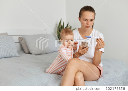 Image of sad woman with sorrow expressing wearing white t shirt sitting on bed with baby daughter, holding smart phone and pregnancy test in hands, being upset of result. 102172856