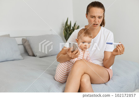 Shocked sad woman wearing white t shirt and shorts sitting on bed with her baby daughter and holding pregnancy test with positive result, disappointed future mother. 102172857