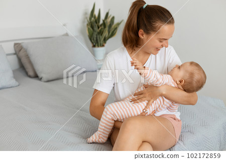 Portrait of happy satisfied attractive woman wearing white t shirt and shorts sitting on bed with her baby daughter, mother playing with her kid, expressing love and gentle. 102172859