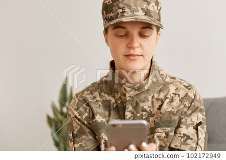 Caucasian young adult female soldier wearing military costume, posing indoor in light room, sitting with smart phone in hands, checking e-mail, having calm concentrated facial expression. Caucasian young adult female soldier wearing military costume, posing indoor in light room, sitting with smart phone in hands, checking e-mail, having calm concentrated facial expression. 102172949