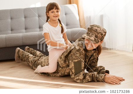 Caucasian female wearing camouflage uniform and cap playing with her daughter after returning home from army, cute daughter sitting on mother's back while mom lying on floor. Caucasian female wearing camouflage uniform and cap playing with her daughter after returning home from army, cute daughter sitting on mother's back while mom lying on floor. 102173286
