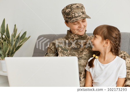 Image of smiling positive woman wearing camouflage uniform and cap, military female returning home after army and spending time with her daughter, sitting and watching something on laptop. Image of smiling positive woman wearing camouflage uniform and cap, military female returning home after army and spending time with her daughter, sitting and watching something on laptop. 102173293