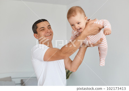 Positive brunette man wearing white casual t shirt holding his infant daughter, posing at home in light room, father lifting hiss daughter in the air, expressing happiness while playing together. Positive brunette man wearing white casual t shirt holding his infant daughter, posing at home in light room, father lifting hiss daughter in the air, expressing happiness while playing together. 102173413