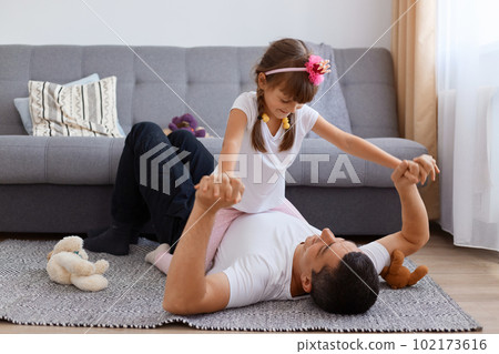 Young adult man lying on floor near sofa and playing with his little kid, wearing white t shirt and trousers, cute girl sitting on father and holding his hands. Young adult man lying on floor near sofa and playing with his little kid, wearing white t shirt and trousers, cute girl sitting on father and holding his hands. 102173616